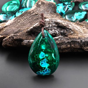 Puede incluir: Un colgante en forma de lágrima hecho de malaquita con un patrón de color verde oscuro y azul. El colgante está colgado de un cordón de cuero marrón.