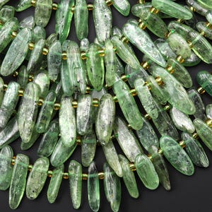 May include: A close-up view of a necklace composed of translucent green gemstone beads. The beads are elongated and faceted, connected with gold-coloured spacers. The necklace has a natural, earthy aesthetic, suitable for jewellery making or personal adornment.