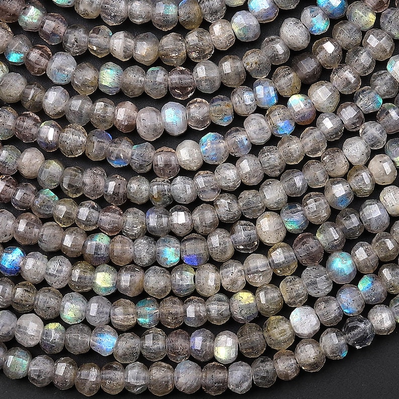 May include: Close-up of strands of faceted labradorite beads. The beads are a mix of gray and translucent with flashes of blue, green, and yellow iridescence. The beads are strung together, creating a textured surface.