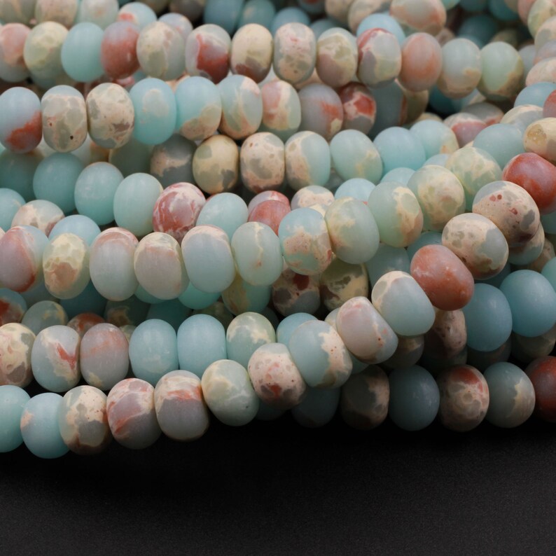 May include: Close-up of a strand of gemstone beads. The beads are a mix of colors, including light blue, beige, and reddish-brown. The beads are oval-shaped and have a mottled appearance. The beads are strung together, creating a necklace or bracelet.