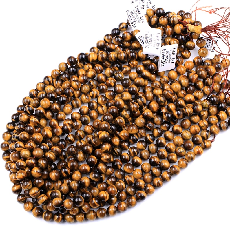 May include: A strand of round, golden brown tiger eye beads. The beads are approximately 6mm in diameter and have a smooth, polished finish. The beads are strung on a brown thread and are ready to be used for jewelry making.