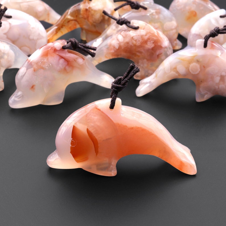 May include: A collection of carved agate dolphin pendants with brown cords. The dolphins are carved in a variety of shades of pink, white, and brown agate.