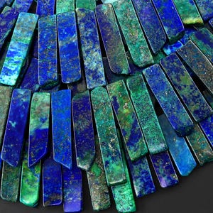 Puede incluir: Una colección de cuentas rectangulares de azurita y malaquita dispuestas en abanico. Cada cuenta presenta una mezcla única de inclusiones minerales en azul real profundo y verde vibrante con motas metálicas de color dorado dispersas.