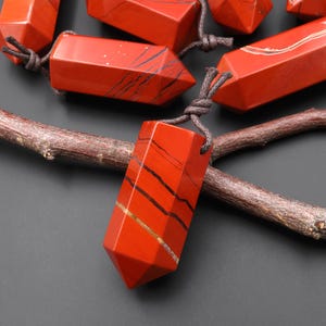 May include: Red jasper gemstone pendants with black and gold veining, strung on brown cords. The pendants are hexagonal and faceted, with a pointed bottom. The gemstones are displayed on a dark background with a wooden branch.