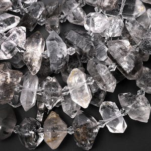 Puede incluir: Primer plano de una hebra de cuentas de cristal de cuarzo transparente con forma puntiaguda y facetada. Las cuentas tienen un aspecto natural y crudo con algunas inclusiones y un ligero tono gris ahumado.