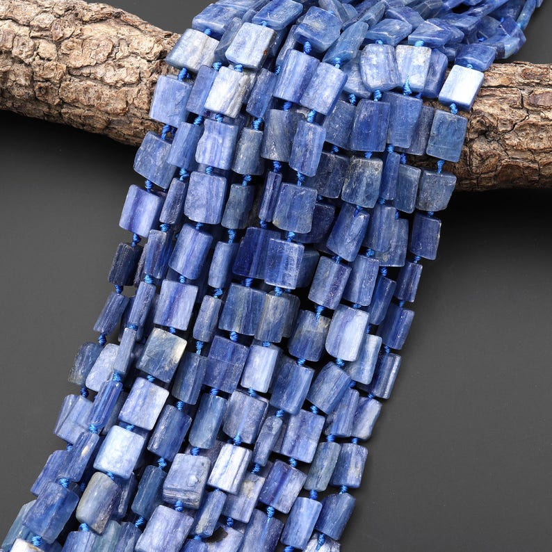 Puede incluir: Hebras de cuentas de cianita azul. Cada cuenta es rectangular con una textura ligeramente rugosa, ensartada con hilo azul. Las cuentas var&iacute;an en tonos de azul, de claro a &iacute;ndigo intenso. El fondo es gris oscuro.