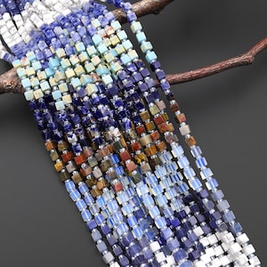 Könnte beinhalten: Stränge aus quadratisch geschliffenen Edelsteinperlen in verschiedenen Farben, darunter Weiß, Blau, Türkis, Braun und Orange. Die Perlen sind mit silberfarbenen Abstandshaltern aufgefädelt. Die Perlen werden vor einem dunkelgrauen Hintergrund und einem braunen Ast präsentiert.