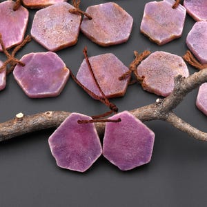 May include: Hexagon-shaped purple gemstone beads with brown stringing. The beads have a natural, textured appearance and vary in shades of purple. A small, brown twig is in the foreground, and the background is a dark gray.
