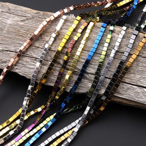 Può includere: Una collezione di perline quadrate in vari colori metallici, tra cui oro, argento, bronzo e tonalità iridescenti. Le perline sono infilate insieme, creando fili che potrebbero essere utilizzati per la creazione di gioielli o altri progetti artigianali. Lo sfondo è una superficie di legno invecchiato.