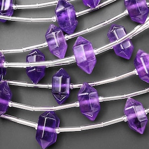 May include: Close-up of amethyst crystal beads strung on silver wire. The faceted, purple crystals are in a variety of shapes, with a polished, translucent appearance. The beads are strung in parallel lines, creating a repeating pattern against a dark background.