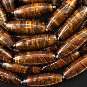 May include: Close-up of a collection of tiger eye gemstone beads. The beads are barrel-shaped, with a polished surface. The colors range from golden brown to dark brown, with striped patterns. The beads are strung together with a white string.