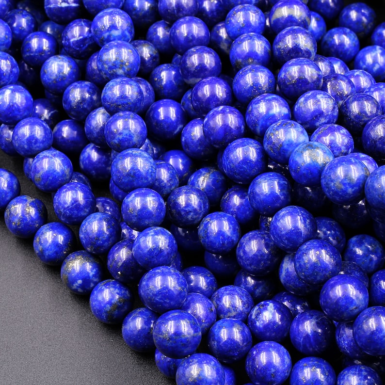 May include: A close-up of a strand of round, blue lapis lazuli beads. The beads are smooth and have a slightly metallic sheen. The beads are arranged in a random pattern, creating a visually appealing texture.