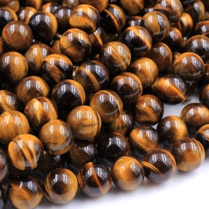 May include: A close-up of a pile of round, polished tiger's eye beads. The beads are a mix of golden brown and black colors with a shimmering, iridescent effect.