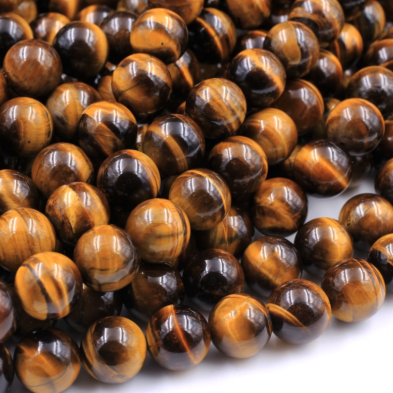 May include: A close-up of a pile of round, polished tiger's eye beads. The beads are a mix of golden brown and black colors with a shimmering, iridescent effect.