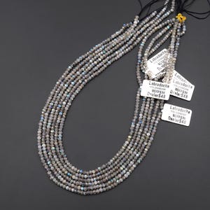 May include: Strands of gray Labradorite gemstone beads. The beads are small, faceted, and strung together on black thread. White tags with the word "Labradorite" and price information are attached.