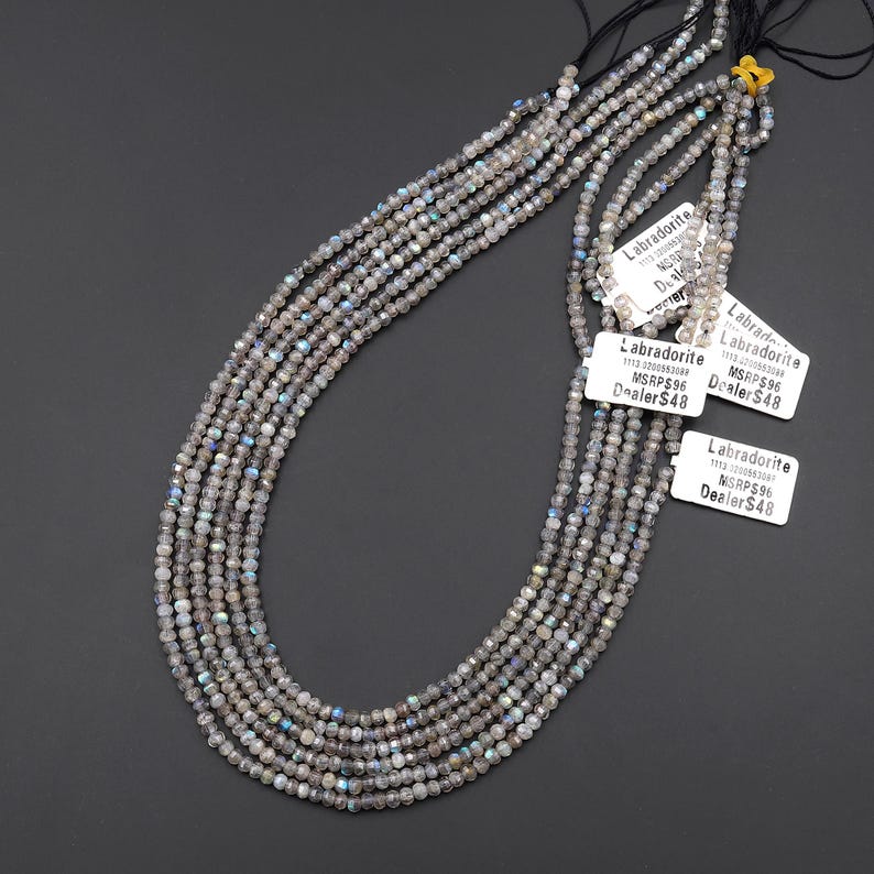 May include: Strands of gray Labradorite gemstone beads. The beads are small, faceted, and strung together on black thread. White tags with the word "Labradorite" and price information are attached.