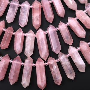 May include: Close-up of a strand of rose quartz crystal beads. The beads are a soft pink color, with variations in shade. Each crystal is a long, pointed shape, strung together with pink string and clear beads. The background is black.