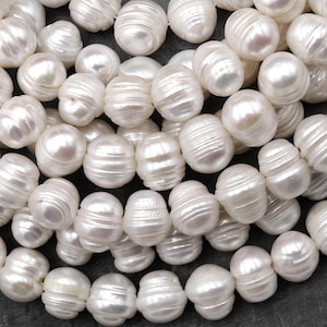 Puede incluir: Primer plano de numerosas perlas barrocas blancas. Las perlas tienen una forma irregular, ovalada, con una superficie estriada y un acabado nacarado brillante. Las perlas están agrupadas, mostrando sus variaciones naturales y texturas únicas.