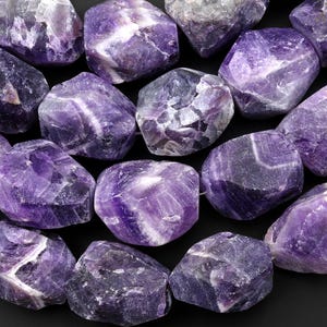 May include: A collection of raw amethyst stones, showcasing a range of purple hues from light lavender to deep violet. The stones have a natural, rough texture and varying sizes, with visible crystalline structures. The arrangement is close-up, highlighting the gemstones' natural beauty.