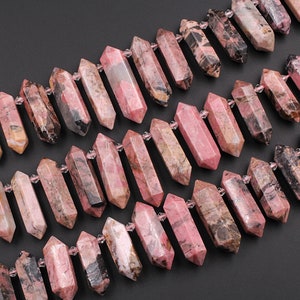 Puede incluir: Una hebra de cuentas de piedras preciosas rosas y negras, cada cuenta tiene una forma rectangular larga y facetada. Las cuentas están enhebradas en un hilo transparente.