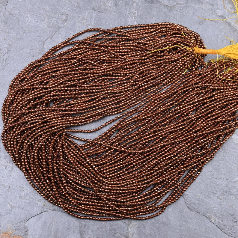 May include: A strand of copper-colored faceted gemstone beads. The beads are small and round, and they have a metallic sheen.