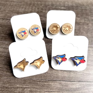 May include: Four pairs of wooden stud earrings on white cards. Two pairs feature the Toronto Blue Jays logo in blue, red, and white. The other two pairs have a circular design and a Blue Jays logo.