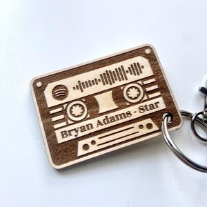 Peut inclure: Un porte-clés en forme de cassette en bois avec le texte "Bryan Adams - Star" gravé. Le porte-clés présente un motif d'onde sonore et un anneau argenté.