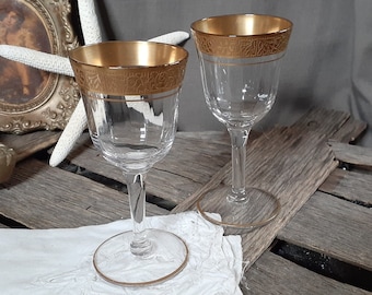 May include: Two clear wine glasses with gold rims and decorative detailing. The glasses sit on a white cloth, with a gold-framed picture and a starfish in the background. The glasses have a classic, elegant design.