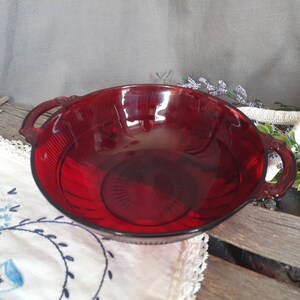 May include: A ruby red glass bowl with two handles. The bowl has a ribbed design on the exterior and a circular pattern on the interior. The bowl is sitting on a white embroidered cloth.