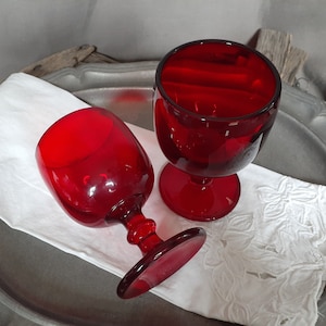 May include: Two ruby red wine glasses with a classic design. The glasses are placed on a white embroidered napkin, set on a silver-toned tray. The stemware has a rounded bowl and a short stem.