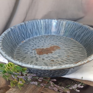 May include: A round, vintage enamelware pan with a blue and white speckled pattern. The pan has a wide rim and a slightly rusted interior. The pan is likely used for cooking or serving food. The overall aesthetic is rustic and charming.
