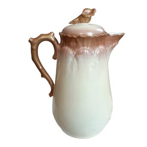 May include: Cream-colored ceramic pitcher with a brown rim and handle. The lid is topped with a decorative brown dog figurine. The pitcher has a bulbous body and a slender spout, suitable for serving beverages.