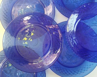 May include: A collection of cobalt blue glass plates featuring a pressed diamond pattern on the outer rim and a central starburst design. The plates are arranged in an overlapping circular formation, showcasing the translucent deep blue glass.