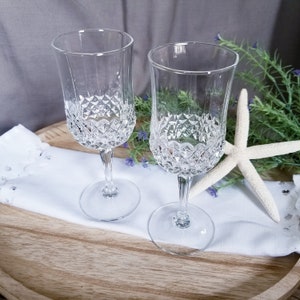 May include: Two clear crystal champagne flutes with diamond-cut detailing on the lower half of the glass. The flutes sit on a white cloth, with a starfish and greenery in the background. The glasses have a classic, elegant design.
