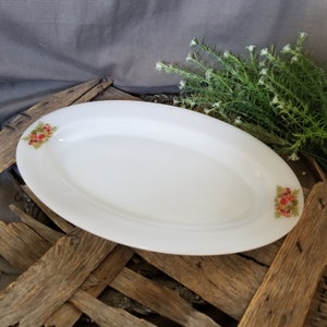 May include: An oval, white serving platter with floral accents. The platter has a smooth surface and is decorated with small clusters of red, yellow, and green flowers on each end. The platter is set on a rustic wooden surface.