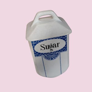 May include: White ceramic sugar dispenser with a blue and white design. The dispenser has a lid with a handle and says "Sugar" in black lettering.