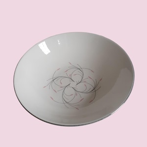 May include: A white ceramic bowl with a silver rim and a floral design. The design features black lines and pink leaf-shaped accents arranged in a symmetrical pattern. The bowl has a smooth, glossy finish.