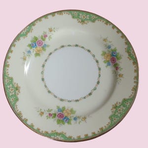 May include: A round, cream-colored plate with a gold rim and floral design. The plate features a central white circle and is decorated with colorful flowers and green accents. The floral pattern includes pink, yellow, and blue flowers.