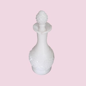 May include: A white glass decanter with a decorative stopper. The decanter has a textured, ribbed neck and a bulbous base with a grape design. The stopper is shaped like a pinecone. The overall height is approximately 10 inches.