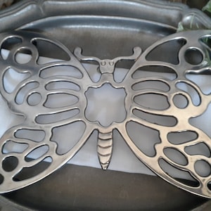 May include: A silver-colored butterfly-shaped trivet, resting on a white cloth napkin and a silver-toned tray. The trivet has an openwork design, with cut-out details forming the butterfly's wings and body. The tray has a decorative edge.