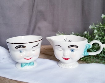 May include: A set of two white ceramic creamer cups with a whimsical face design. The cups feature a winking face with a blue eye and a red mouth. The cups are decorated with the text "Baileys" and a blue bow.