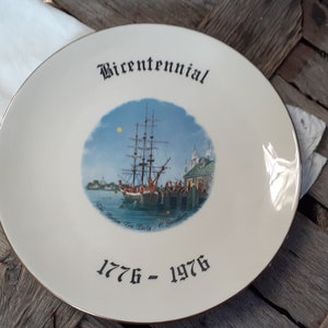 May include: A round, off-white ceramic commemorative plate featuring a central illustration of a tall ship docked at a harbor. The word Bicentennial is printed in black above the image, with the years 1776 - 1976 printed in black below the image.