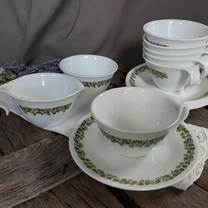 May include: A set of white ceramic cups, saucers, and bowls with a green floral pattern. The cups have handles and are stacked. The saucers and bowls are also white with the same green floral design. The items are arranged on a wooden surface.