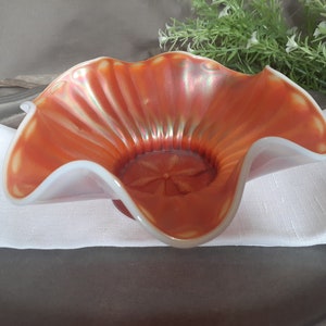 May include: An orange iridescent glass bowl with a ruffled edge. The bowl has a white rim and a starburst design in the center. The bowl is approximately 6 inches in diameter.