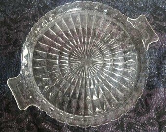 May include: Clear glass serving tray with a circular design and two handles. The tray features a textured pattern radiating from the center, with a scalloped edge. The handles are rectangular with a decorative shape.