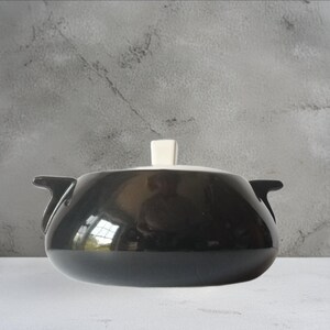 May include: A glossy black ceramic casserole dish with a rounded body, two side handles, and a white lid featuring a square-shaped knob handle. The surface of the black pot is reflective, showing a faint light reflection on its curved exterior.
