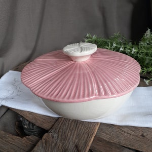 May include: A ceramic lidded bowl featuring a cream-colored base with a subtle crackle glaze finish. The lid is a soft pink color with a speckled texture and a radial ribbed pattern, topped with a white, flower-shaped handle knob.