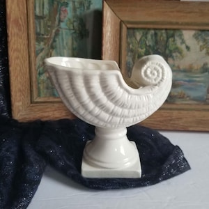 May include: A white ceramic vessel shaped like a nautilus shell, featuring a ribbed exterior texture and a prominent spiral detail at one end. The shell rests on a square pedestal base, creating a raised, open-top container with a glossy finish.