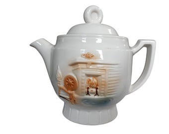May include: White ceramic teapot with a lid and handle. The teapot features a raised relief design of a spinning wheel and a sheep in a barn, with tan and brown accents. The spout and handle are smooth.