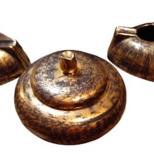 May include: Three-piece ashtray set with a metallic gold and brown finish. The set includes two triangular ashtrays and a round, lidded container. The ashtrays have a classic design with a recessed area for holding cigarettes.